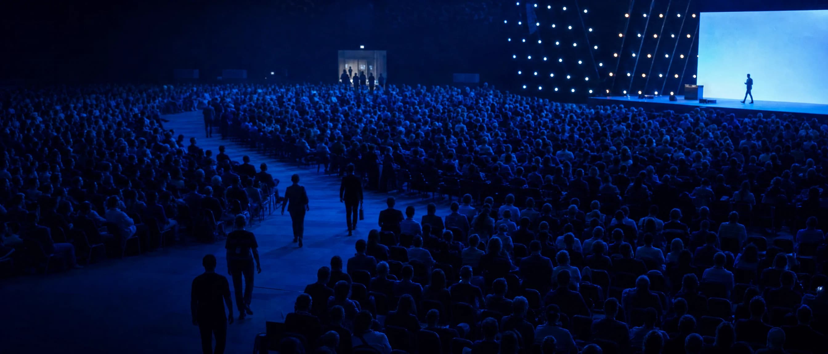 A conference hall in blue light, a speaker on stage, the audience seated